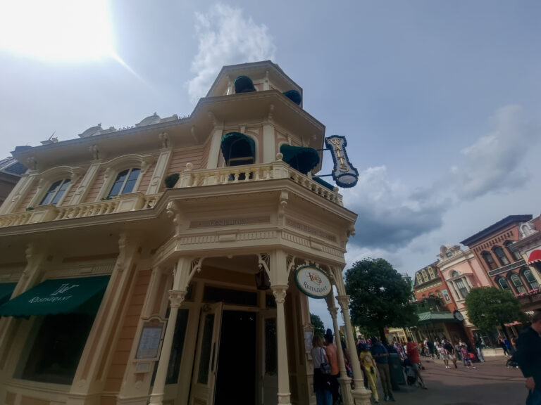 Walt’s – an American Restaurant sits on the corner of Main Street with ornate balconies, green awnings, and guests walking by on a sunny afternoon.