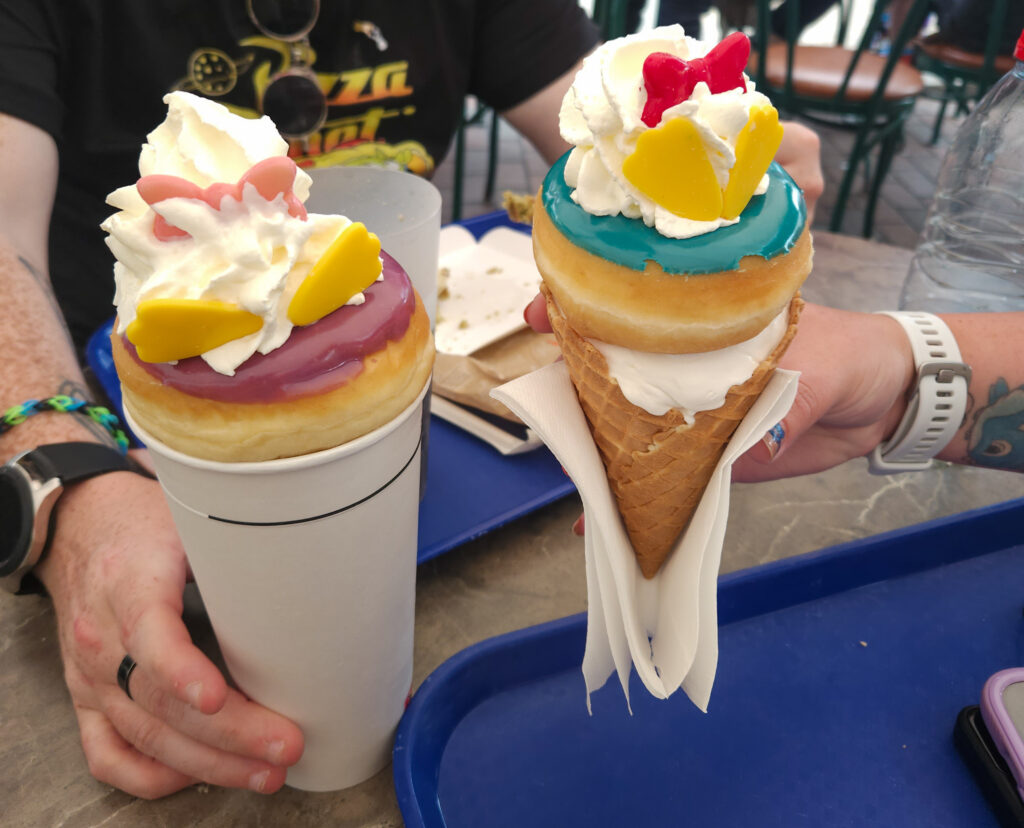 Two character-themed donut desserts—one served in a cup and one in a cone—decorated with colored icing, whipped cream, and candy pieces shaped like bows and shoes. These are whimsical Disneyland Paris snacks perfect for photo ops.