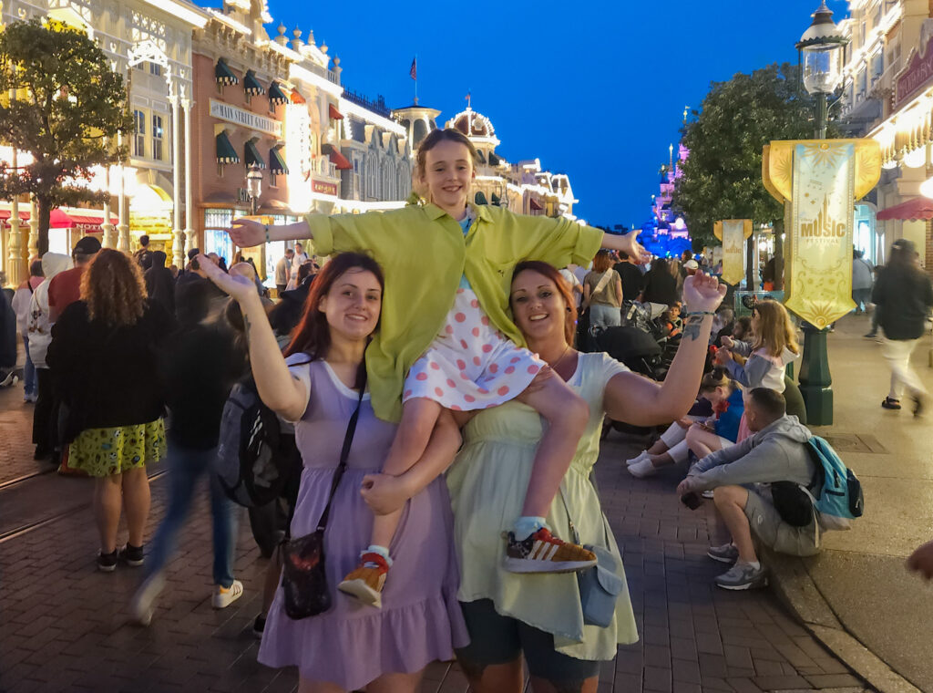 Three guests dressed in pastel outfits pose joyfully on Main Street U.S.A. at night, with one child lifted high on the shoulders of two smiling adults. The street sparkles with lights, and Sleeping Beauty Castle glows in the background.