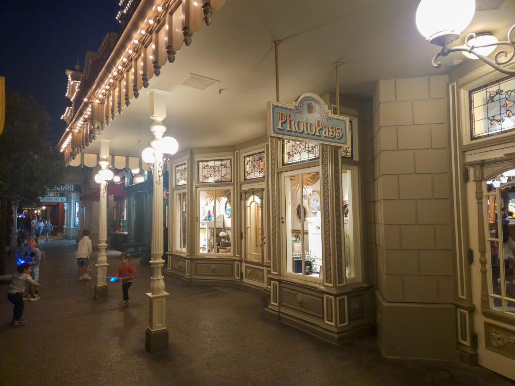 The illuminated storefront of the Emporium at night, glowing with vintage-style lamps and showcasing Disney merchandise through its arched display windows.