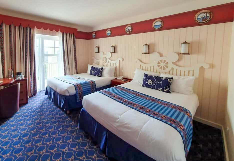 Cheerful nautical-themed hotel room with two beds, ship wheel headboards, blue and red accents, and porthole-style framed artwork along the top wall.