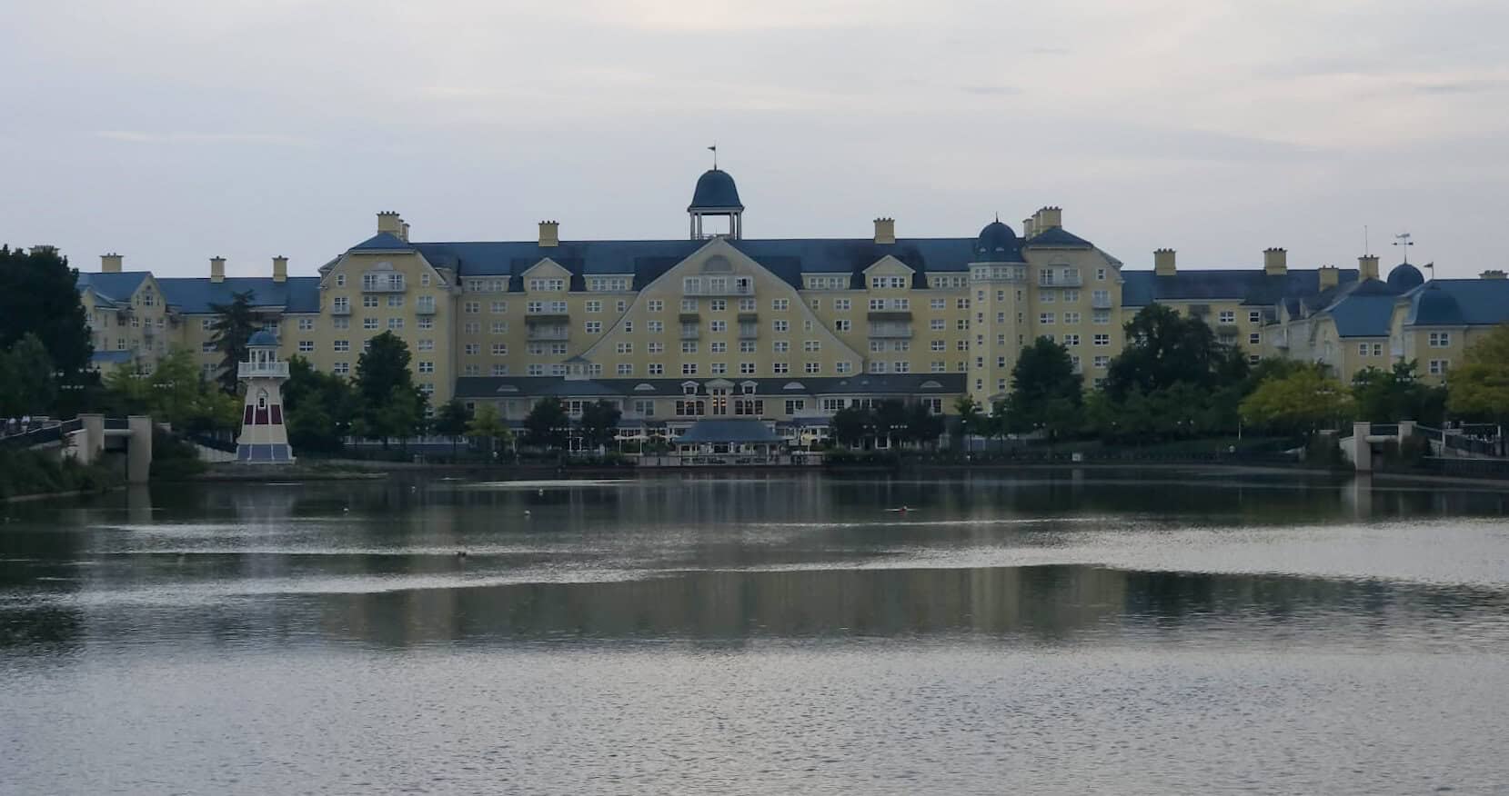 Elegant lakeside view of Disney’s Newport Bay Club, a sprawling yellow hotel with blue rooftops and a lighthouse near the water, reflecting a nautical theme.