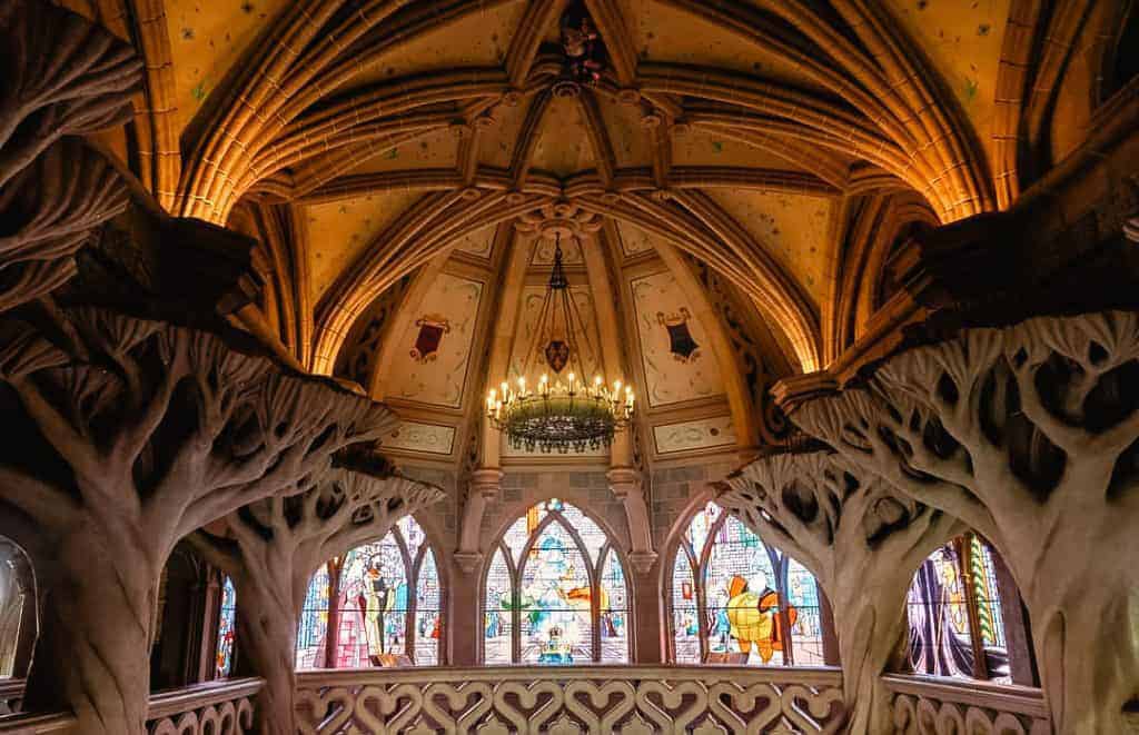 A beautifully lit stained-glass gallery under vaulted stone arches, depicting scenes from Sleeping Beauty with carved tree pillars and chandeliers.