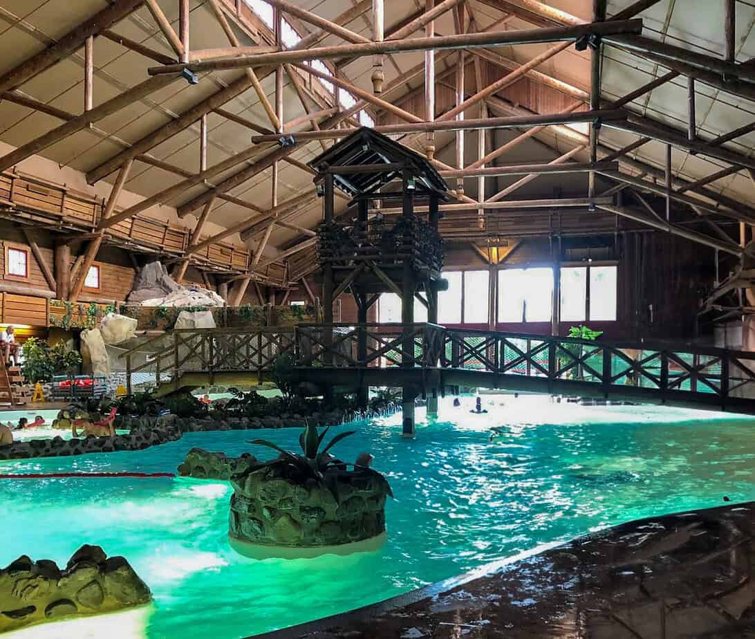 Rustic indoor water park with a log cabin theme, featuring a wooden bridge, artificial rocks, and glowing turquoise water—one of the most adventurous disneyland paris hotels with pools.