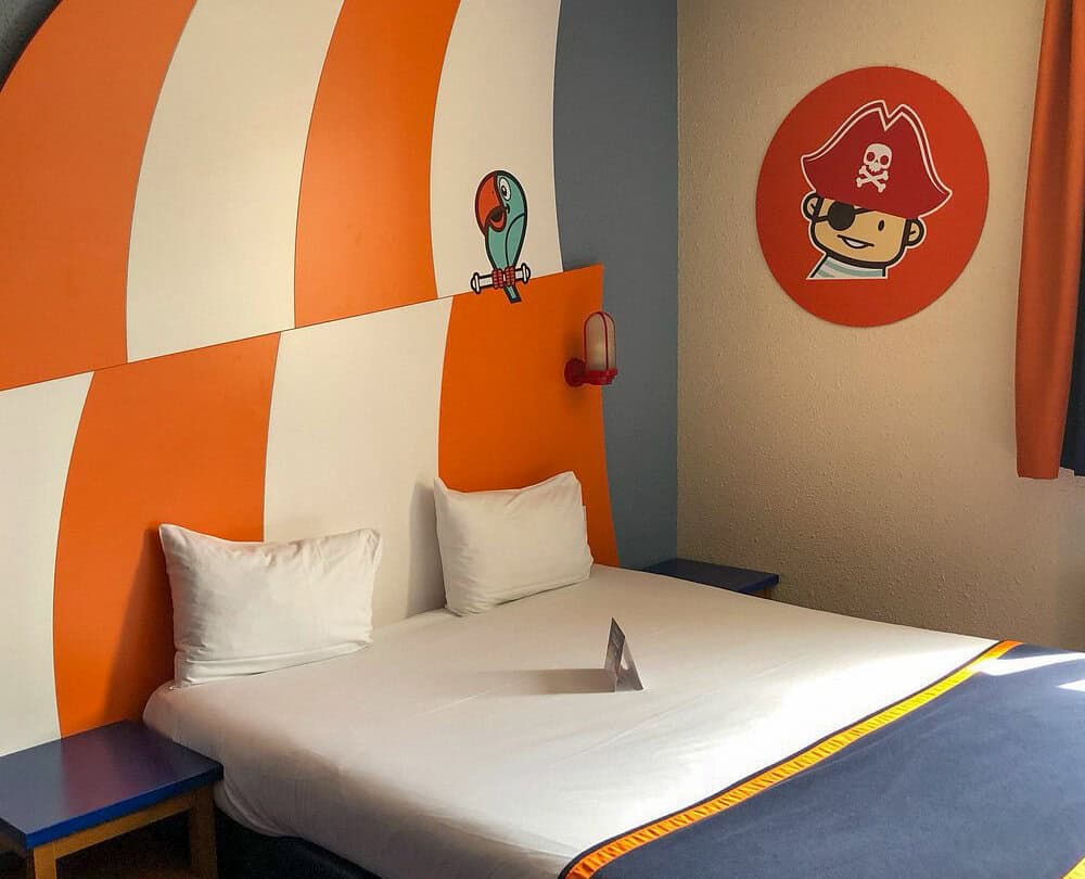 Bright, playful hotel room with a nautical pirate theme, featuring cartoon artwork of a parrot and pirate above the bed on orange and white striped walls.