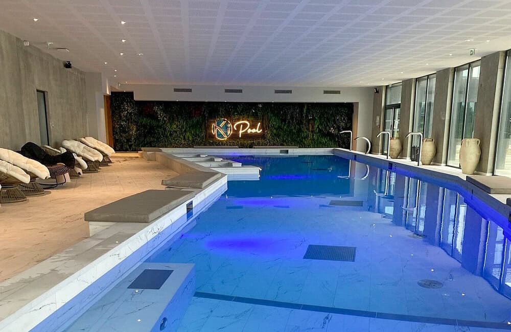 Luxurious indoor pool at a modern hotel spa, featuring lounge chairs, marble floors, and soft blue lighting under a sleek, minimal ceiling.