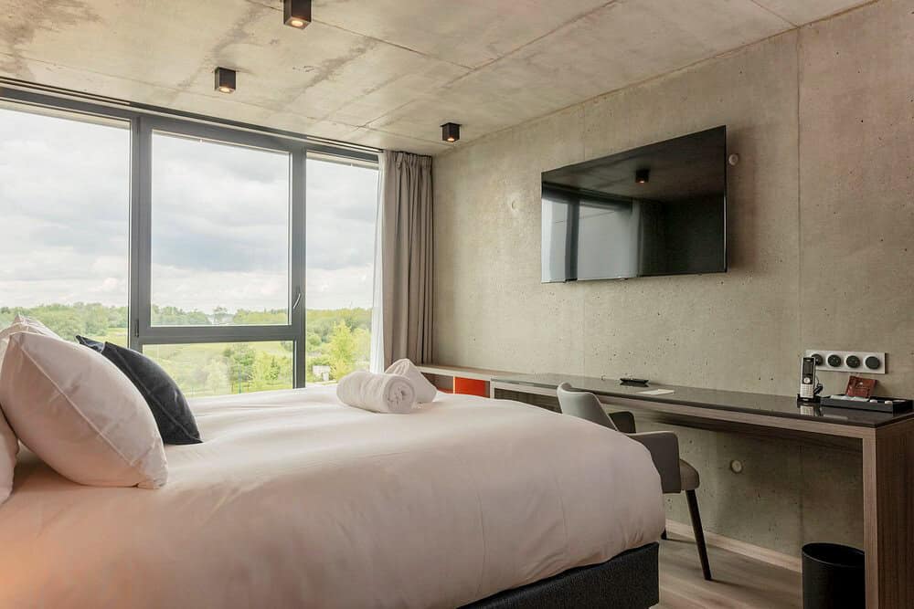 Sleek and modern hotel room with concrete walls, large windows overlooking greenery, a mounted flat-screen TV, and a tidy work desk.