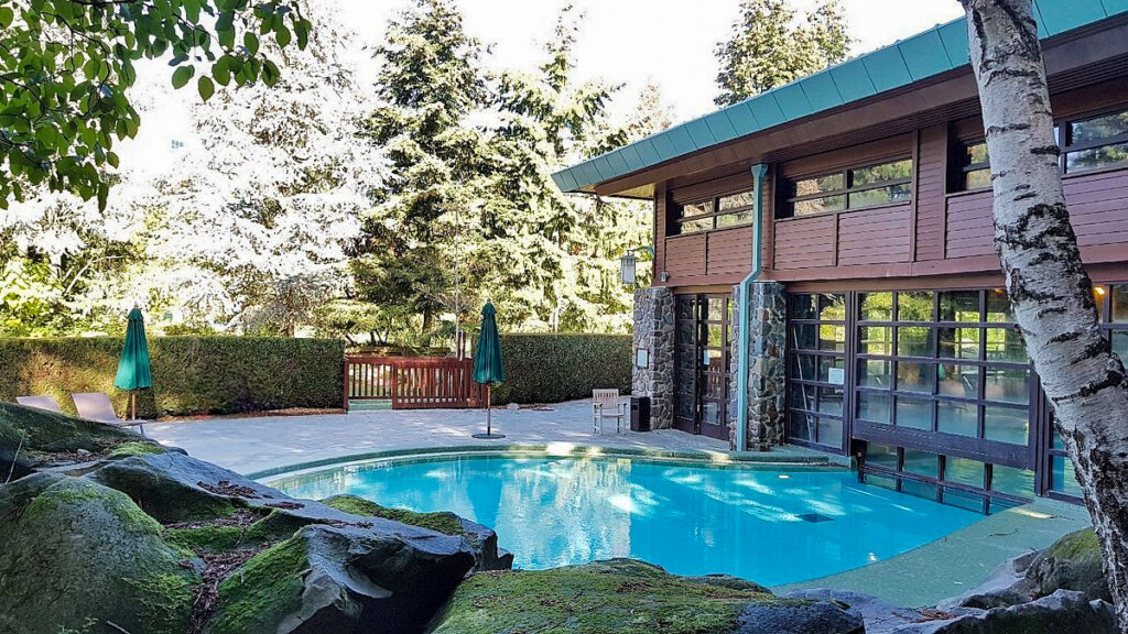 Tranquil outdoor pool area surrounded by greenery and lodge-style architecture, with lounge chairs and umbrellas on the deck.
