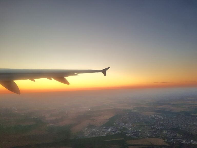 The wing of a plane in mid-air captured during sunrise, with golden light casting over farmlands and towns below.