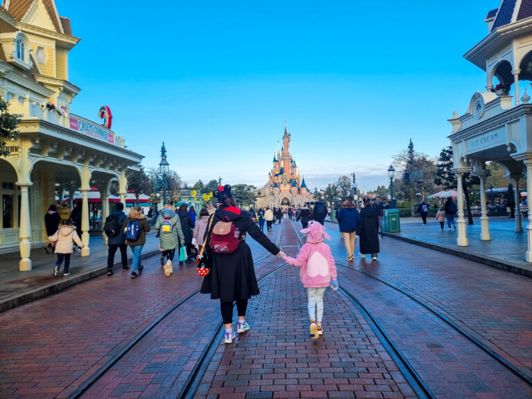 A parent and child hold hands while walking down Main Street, U.S.A. at Disneyland Paris, with the Sleeping Beauty Castle in the distance and the brick path glistening slightly after a light Disneyland Paris rain.