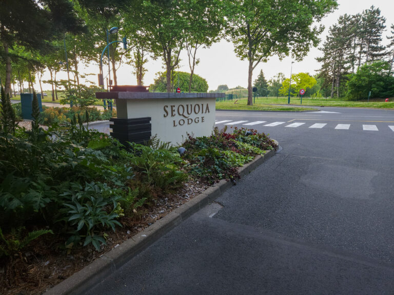 Concrete roadside sign reading “SEQUOIA LODGE” near a crosswalk, surrounded by landscaped greenery and trees, indicating the entrance to one of the Disneyland Paris on site hotels.