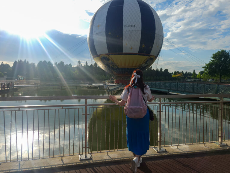 A woman wearing a pink backpack stands at a waterfront railing, gazing at a massive tethered balloon adorned with stars, under golden morning sunlight. A peaceful start to a Disneyland Paris solo day.