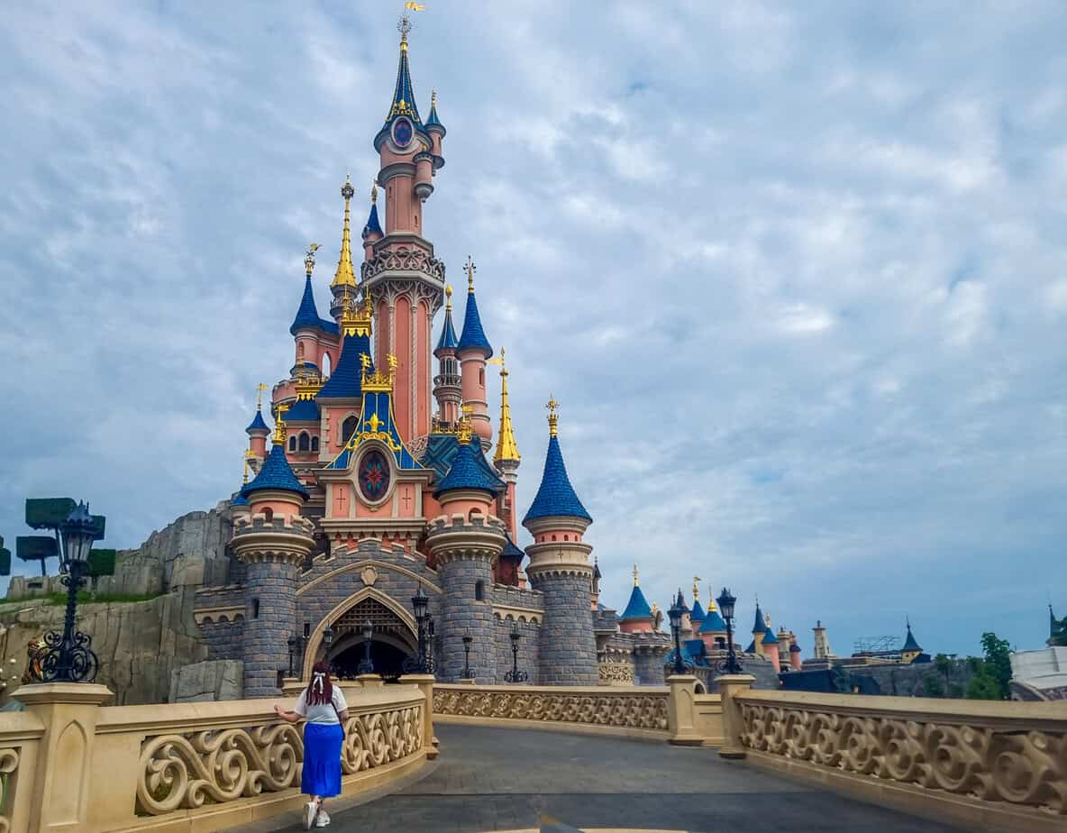 A woman walks toward Sleeping Beauty Castle at Disneyland Paris, with vibrant blue rooftops and golden spires glowing under a cloudy sky.