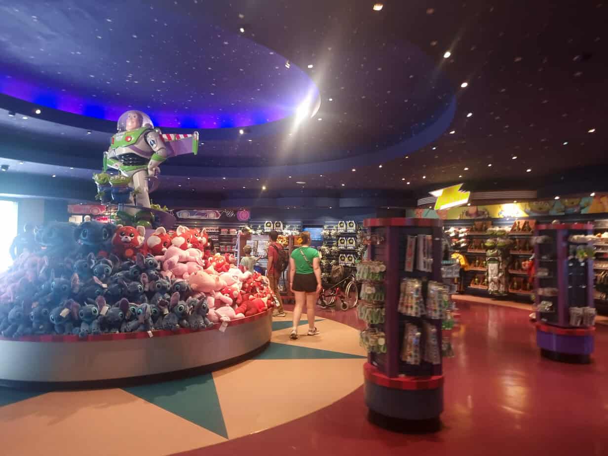 Inside the World of Disney store, a central display features a giant Buzz Lightyear figure atop plush mountains of Stitch and other Disney characters, surrounded by merchandise and shoppers—part of the immersive Disneyland Paris Toy Story experience.