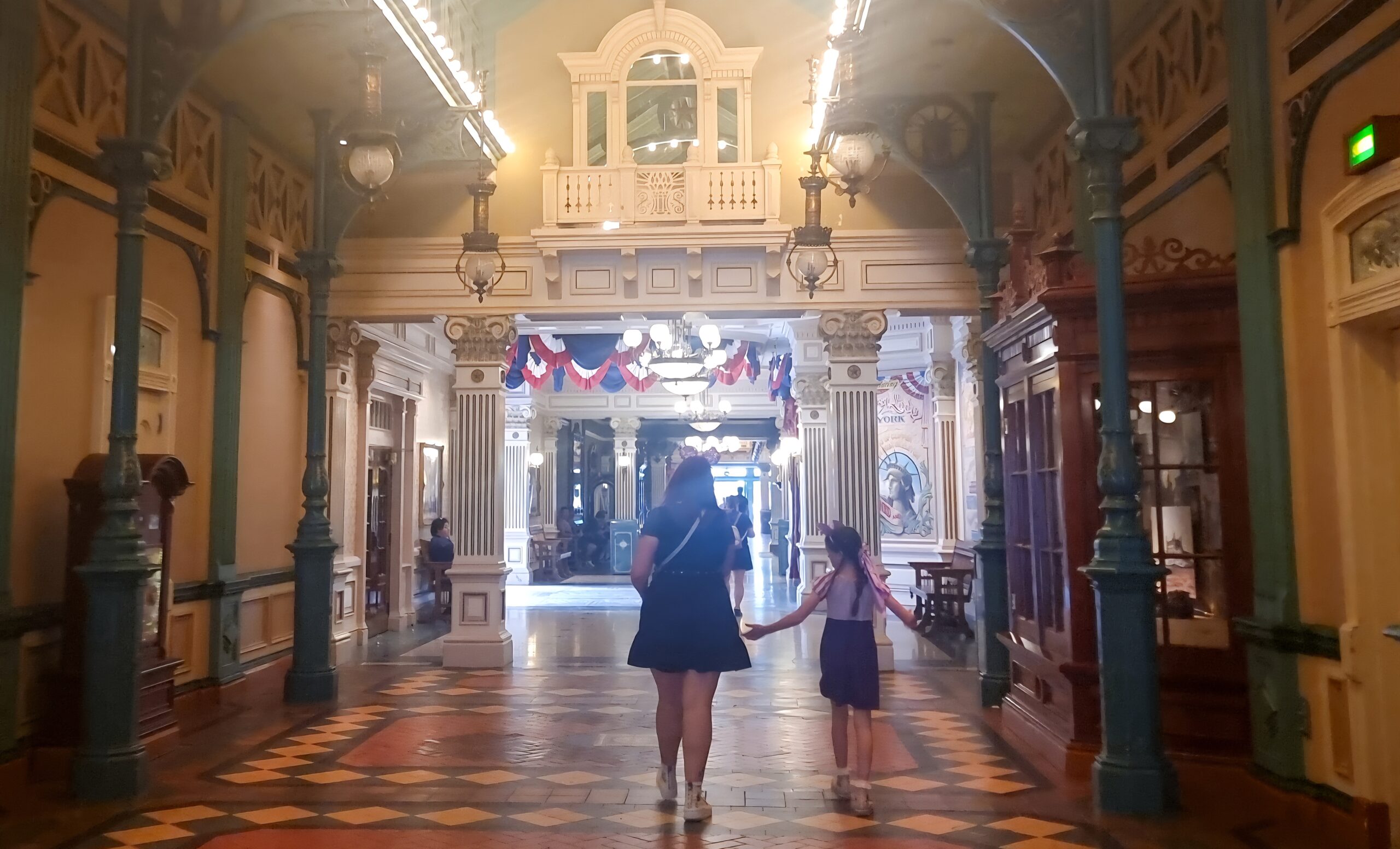 A woman and child walk hand-in-hand through an elegant indoor arcade with vintage lanterns, tiled floors, and patriotic bunting—capturing a quieter side of Disneyland Paris photo spots.