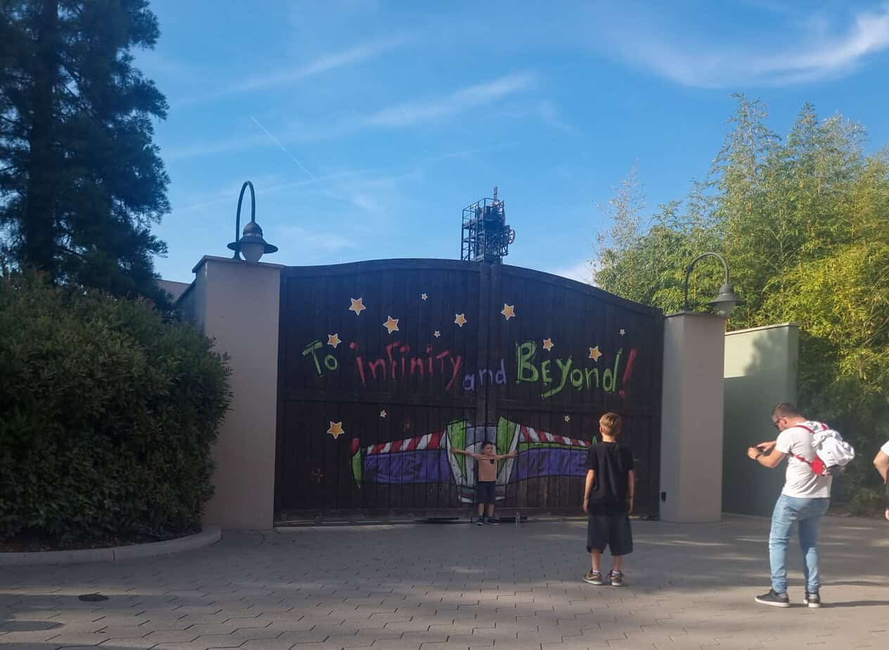 Children pose in front of a large wooden gate painted with Buzz Lightyear wings and the phrase “To infinity and beyond!” set against a bright blue sky.