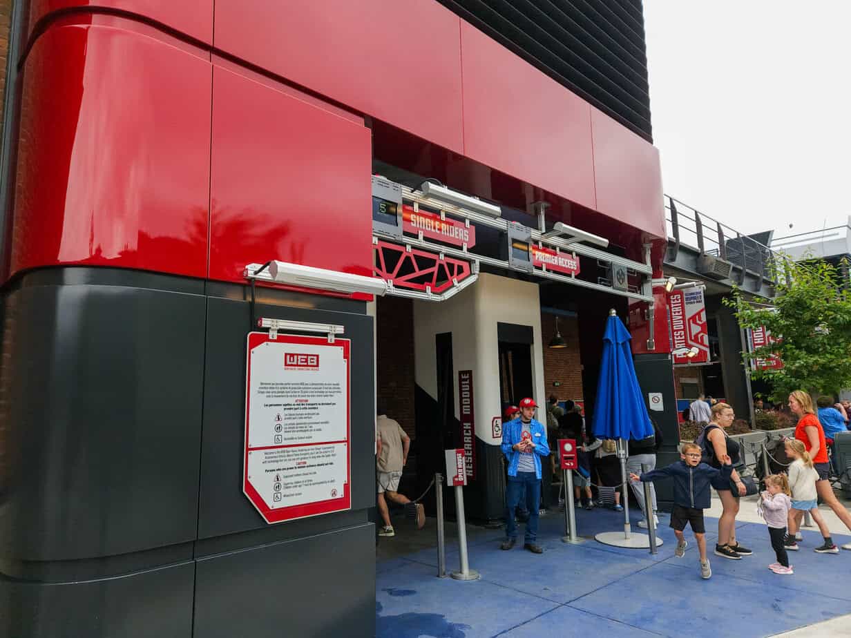 The entrance to the WEB Slingers attraction features red and gray industrial styling, with signage for single riders and Premier Access, as families walk past.