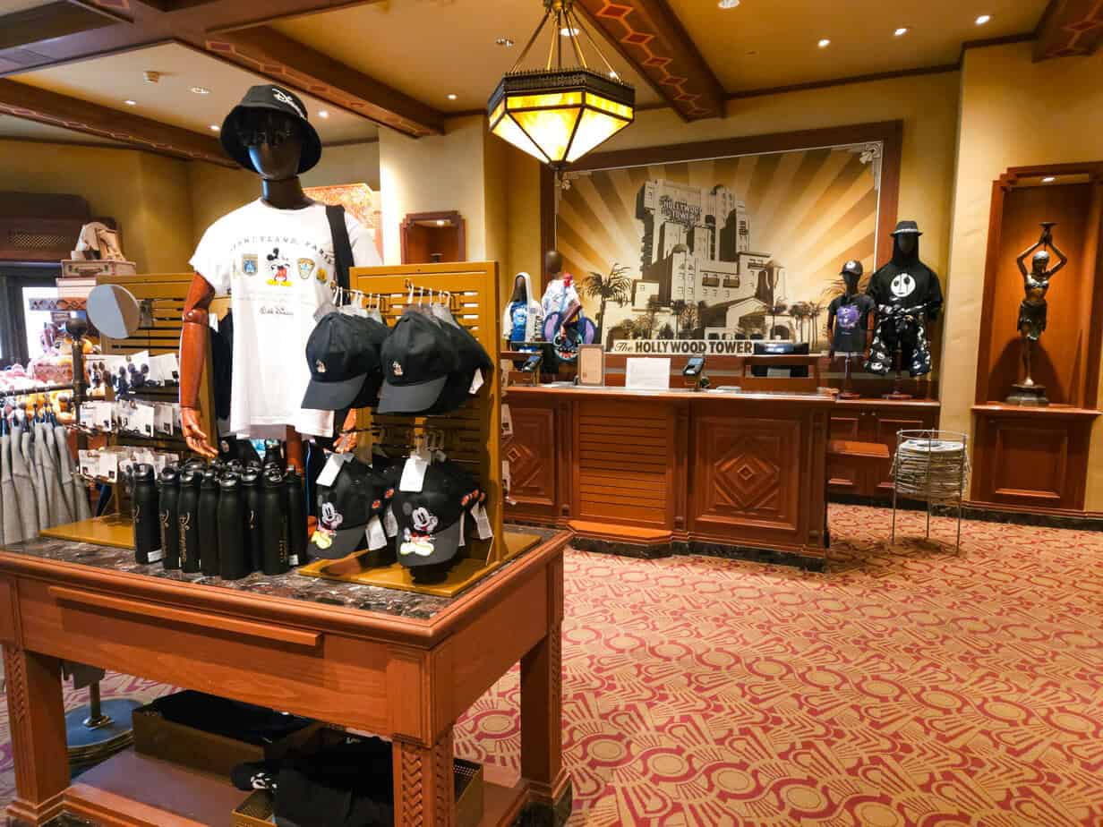 Inside a Disney-themed gift shop, black Mickey Mouse hats, water bottles, and apparel are neatly arranged under a Hollywood Tower mural.
