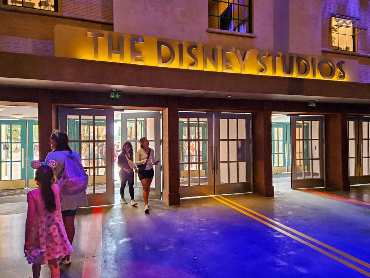 Guests walk through the entrance of The Disney Studios building, where warm yellow lighting highlights the sign above a row of glass doors.