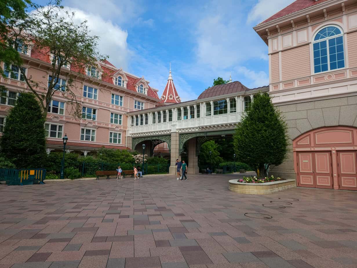 A wide view of the Disneyland Hotel’s pink exterior and covered walkway, with a few people strolling across the quiet plaza.