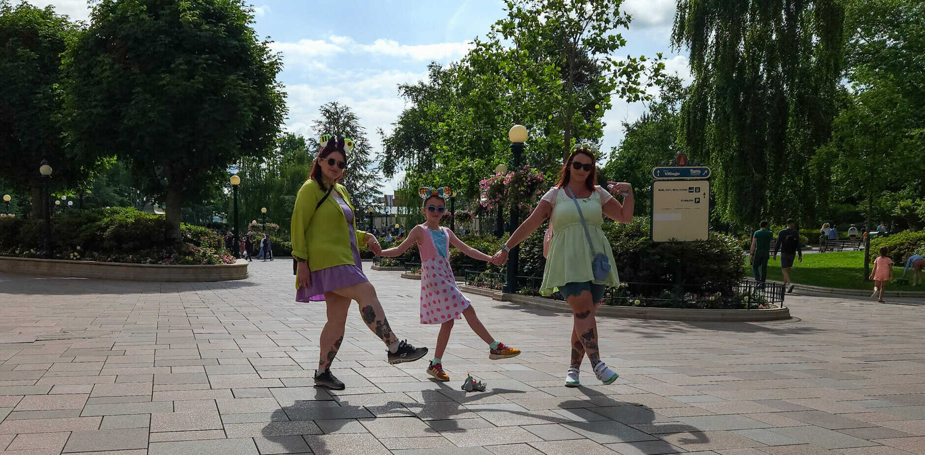 Three people strike a playful pose while holding hands in the middle of a paved walkway lined with flowers and trees on a sunny day.