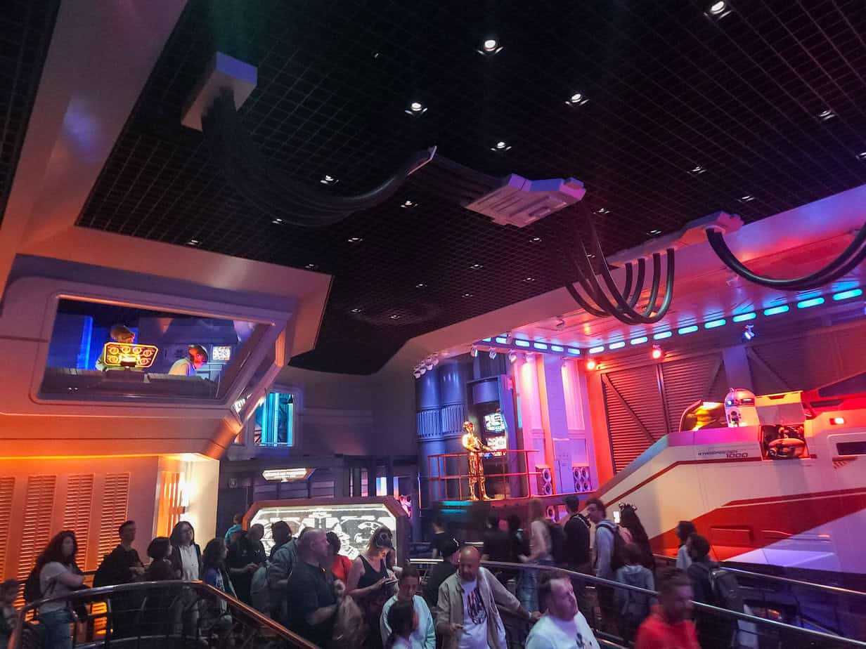 Park guests navigate the indoor Star Tours line, passing animatronic characters including C-3PO. The room glows with red, orange, and blue lighting, adding to the immersive spacecraft hangar theme.