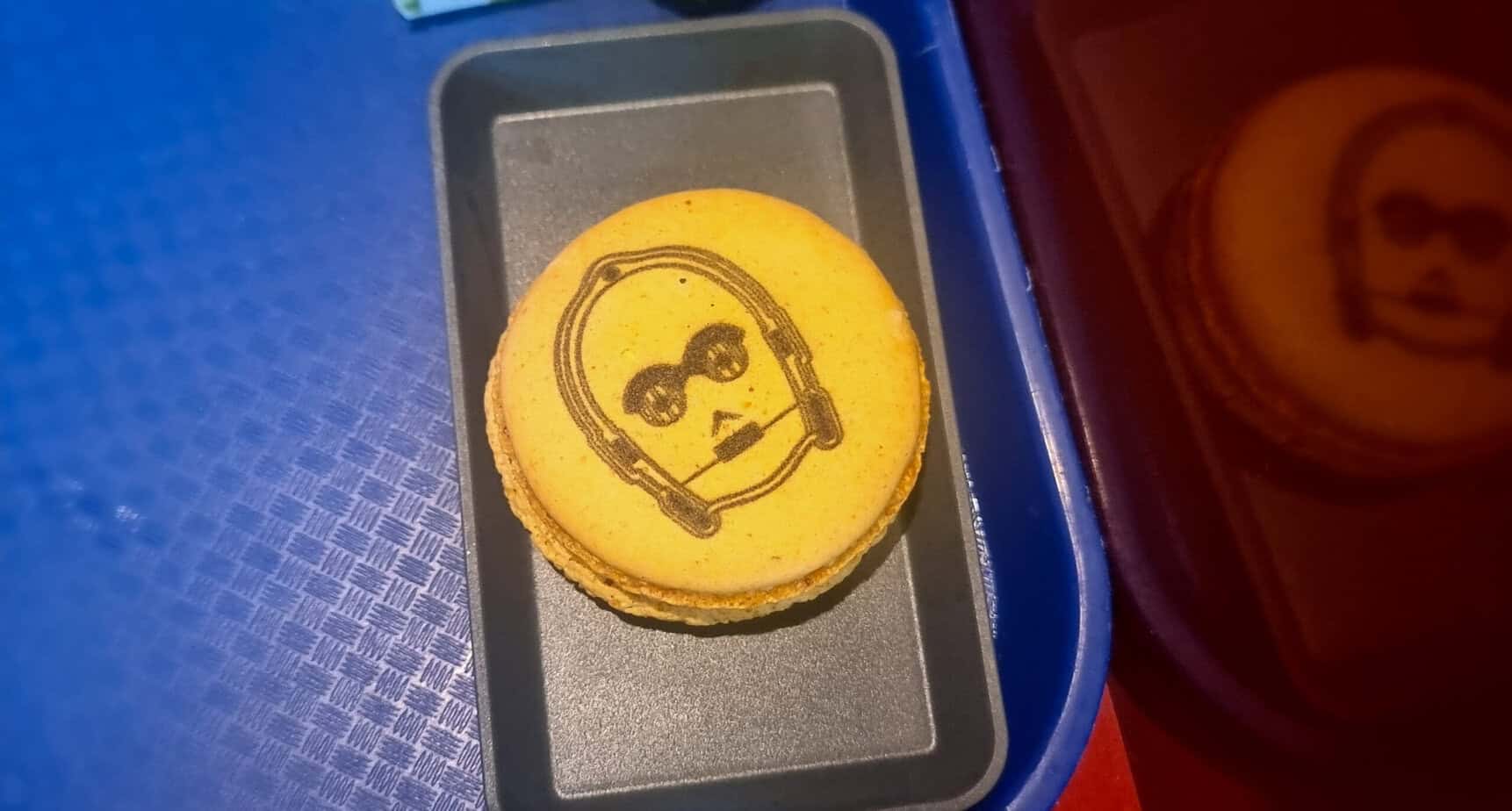 A yellow macaron featuring a black print of C-3PO’s face sits on a black serving tray. This themed dessert is a whimsical Star Wars treat offered at Disneyland Paris.