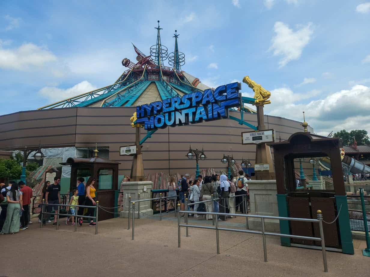 Outside the Hyperspace Mountain attraction, visitors queue near a large retro-futuristic building topped with teal and bronze detailing. The bold ride sign arches above the crowd under a clear blue sky.