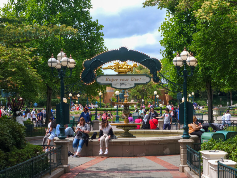 A welcoming fountain area near the entrance of Disneyland Paris, framed by an archway reading “Enjoy your Day!” as visitors relax and stroll.