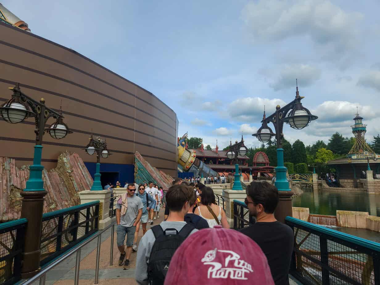 Guests walk along a themed bridge toward the Hyperspace Mountain attraction in Disneyland Paris, surrounded by steampunk-style lampposts and water features.