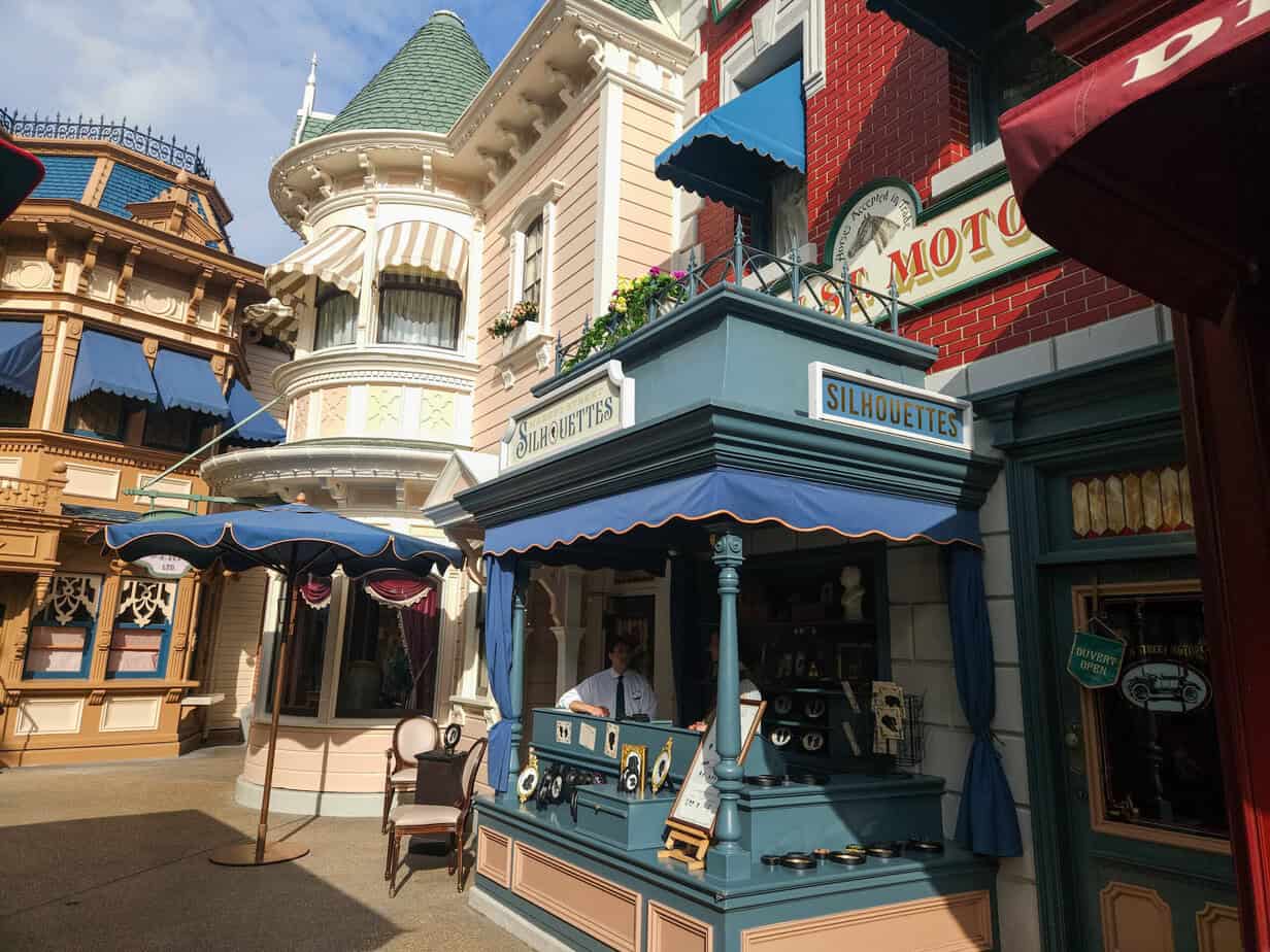 A small street-side stand labeled “Silhouettes” in a vintage-style Disney storefront, with a cast member inside and charming architectural details around.