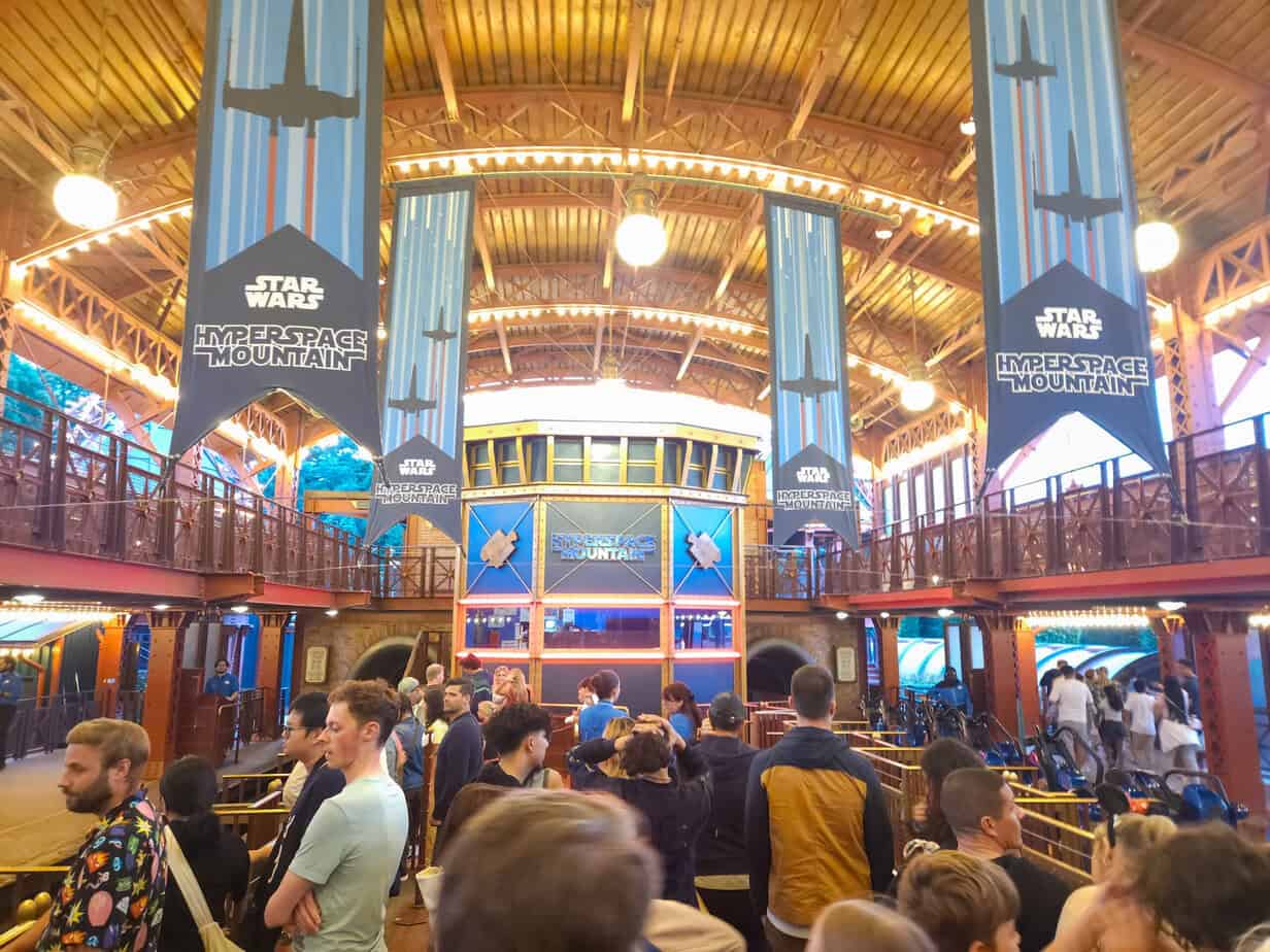 Guests wait inside the Hyperspace Mountain queue area, surrounded by tall “Star Wars Hyperspace Mountain” banners and a vaulted wooden ceiling. The space blends steampunk with sci-fi as riders approach the launch platform.