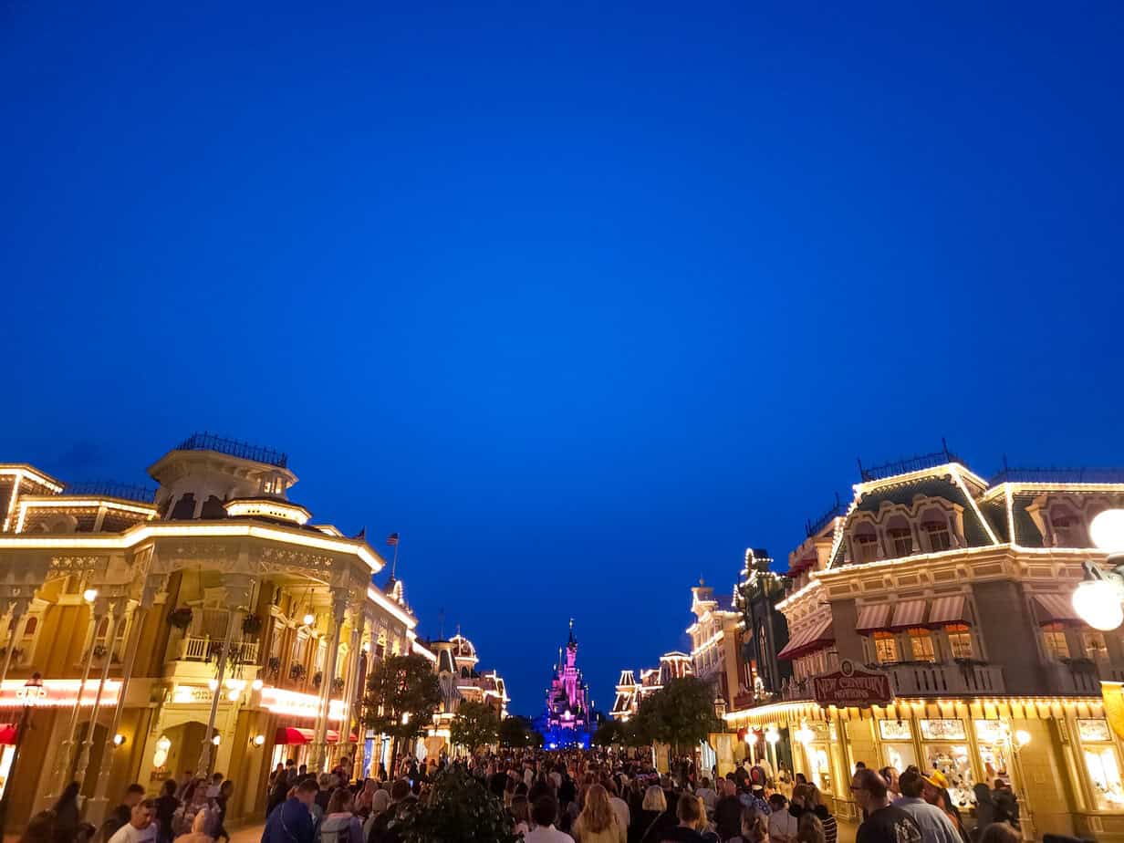 A view of Main Street U.S.A. at Disneyland Paris lit up at night, with the Sleeping Beauty Castle glowing in the distance.
