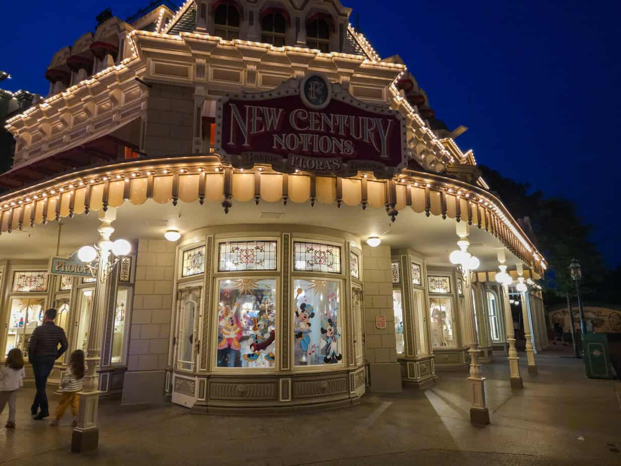 A warmly lit corner shop at night with a sign reading “New Century Notions Featuring Flora’s,” showcasing Disney-themed window displays and a family walking past.
