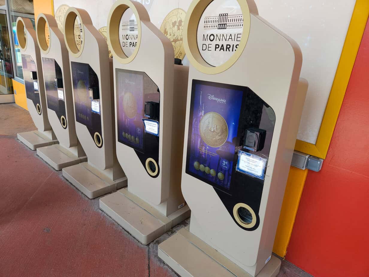 A row of Monnaie de Paris souvenir coin machines at Disneyland Paris, each offering commemorative Disney-themed coins for €3, styled in cream and gold.
