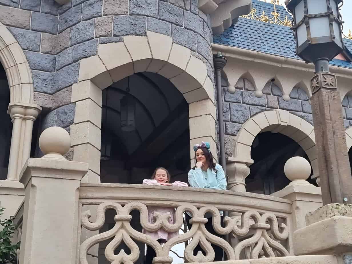 Two people lean on a stone railing at Sleeping Beauty Castle, smiling while surrounded by gothic arches and textured castle walls.