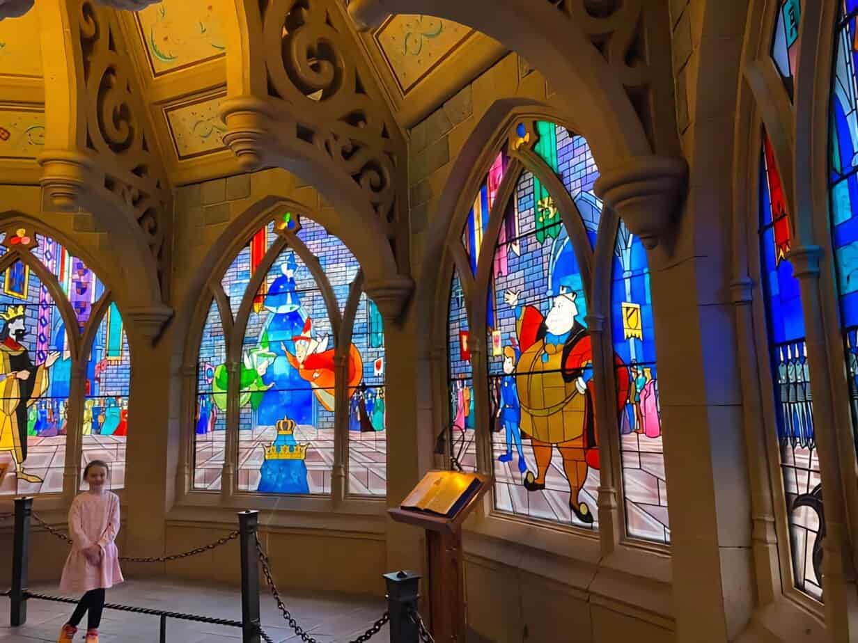 A child stands in a glowing corridor of stained glass windows inside Sleeping Beauty Castle, featuring colorful scenes from the fairytale.