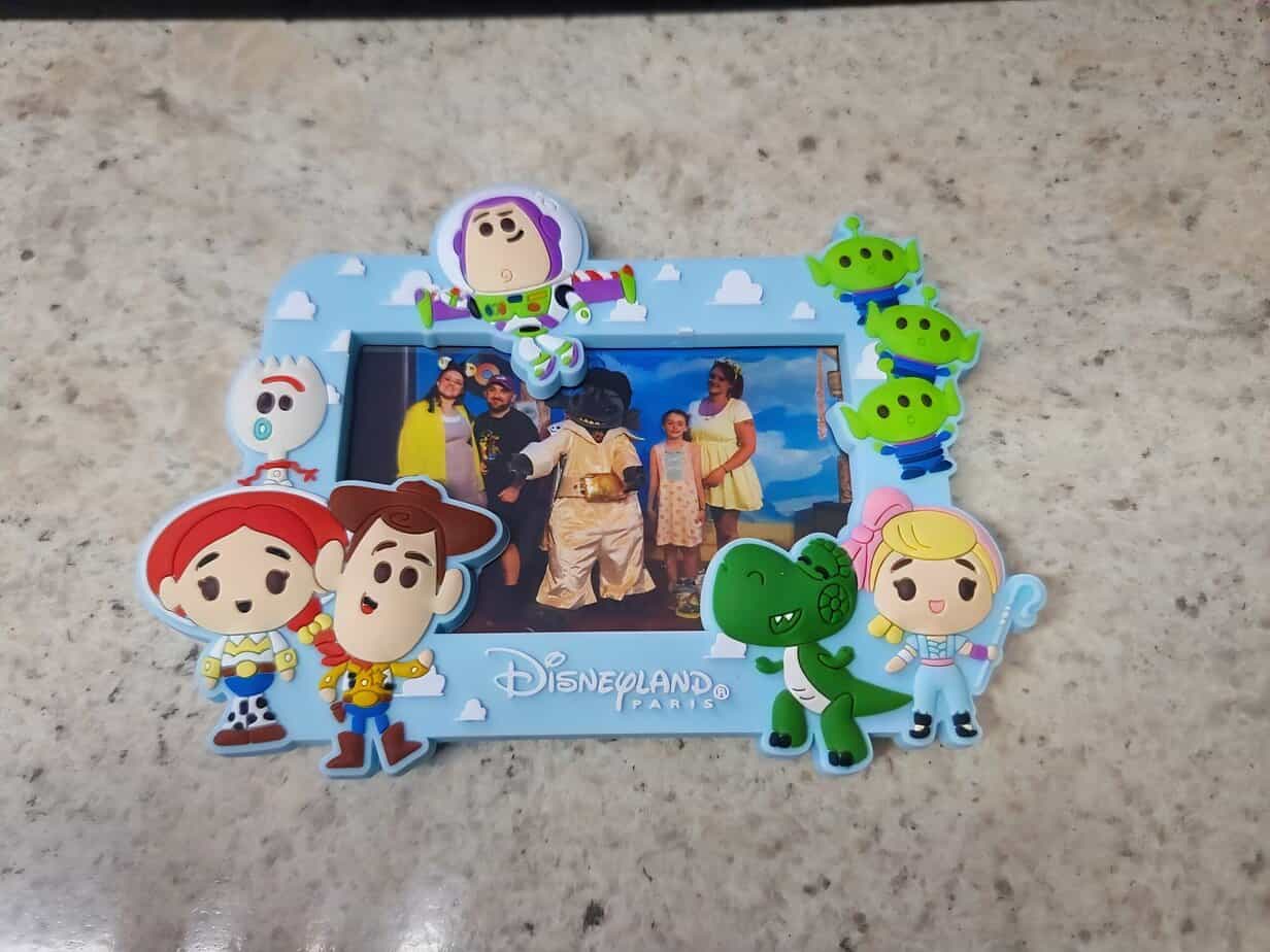 A Disneyland Paris photo frame decorated with cartoon-style Toy Story characters like Woody, Buzz Lightyear, Jessie, and the aliens, surrounding a family photo with Disney cast members inside.