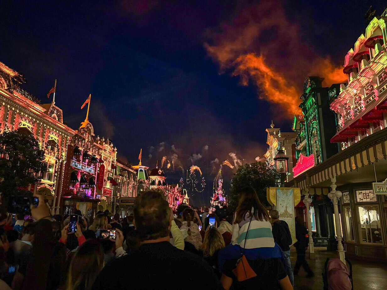 A nighttime crowd gathers on Main Street, U.S.A. at Disneyland Paris, watching a fireworks show above the illuminated Sleeping Beauty Castle, with vibrant lights and glowing orange clouds overhead.