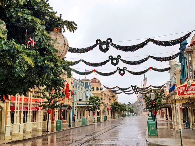 Main Street in Disneyland Paris decorated for the holidays with hanging garlands featuring Mickey-shaped wreaths and colorful ornaments. The view leads to Sleeping Beauty Castle in the distance, capturing the festive charm of Christmas at Disneyland Paris.