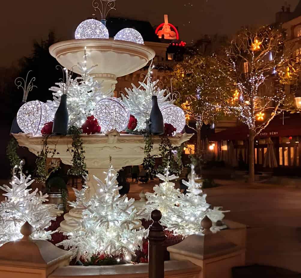 Elegant nighttime holiday display in a fountain with white trees, glowing globe ornaments, and bottles resembling champagne surrounded by twinkling lights.