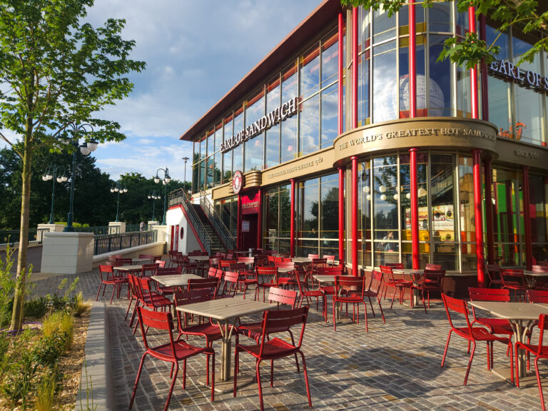Bright morning view of Earl of Sandwich with red patio tables and a glass-fronted building surrounded by trees and lampposts in Disneyland Paris' Disney Village.