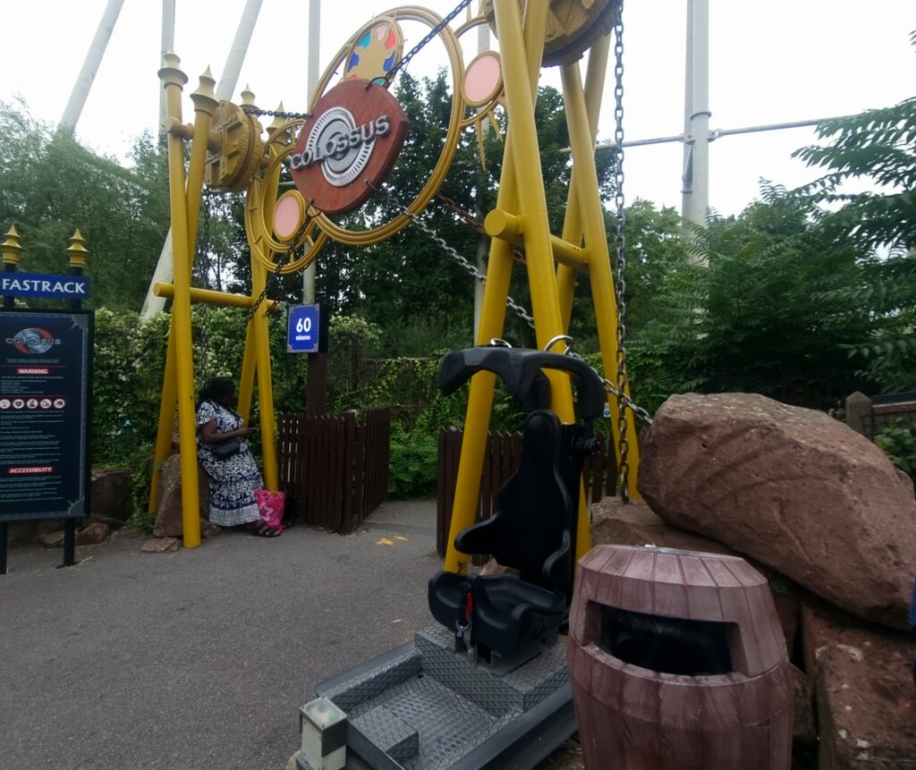A black test seat with harness is positioned at the entrance to Colossus, alongside a themed archway and signage showing a 60-minute wait. These test seats are essential for managing expectations around fit, especially for those concerned about plus size at Thorpe Park.