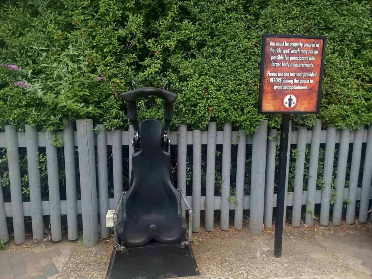 A standalone black test seat is mounted in front of a leafy hedge with a sign advising guests with larger body measurements to check fit before queuing. This is a common practice supporting guest comfort, especially regarding plus size at Thorpe Park.