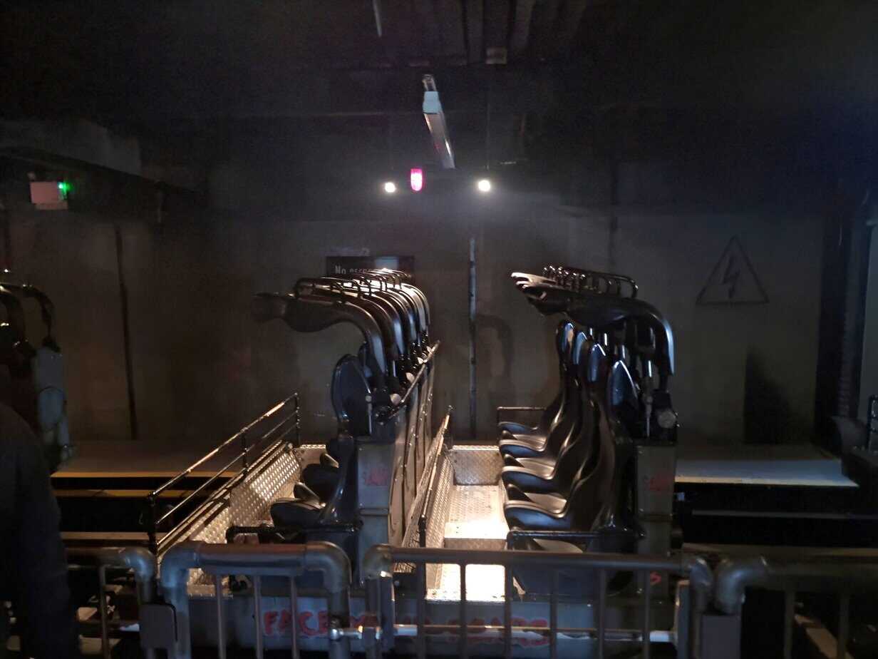 An empty rollercoaster train with sleek black seats sits in a dimly lit loading station with warning signs and grated flooring, ready for the next set of riders.