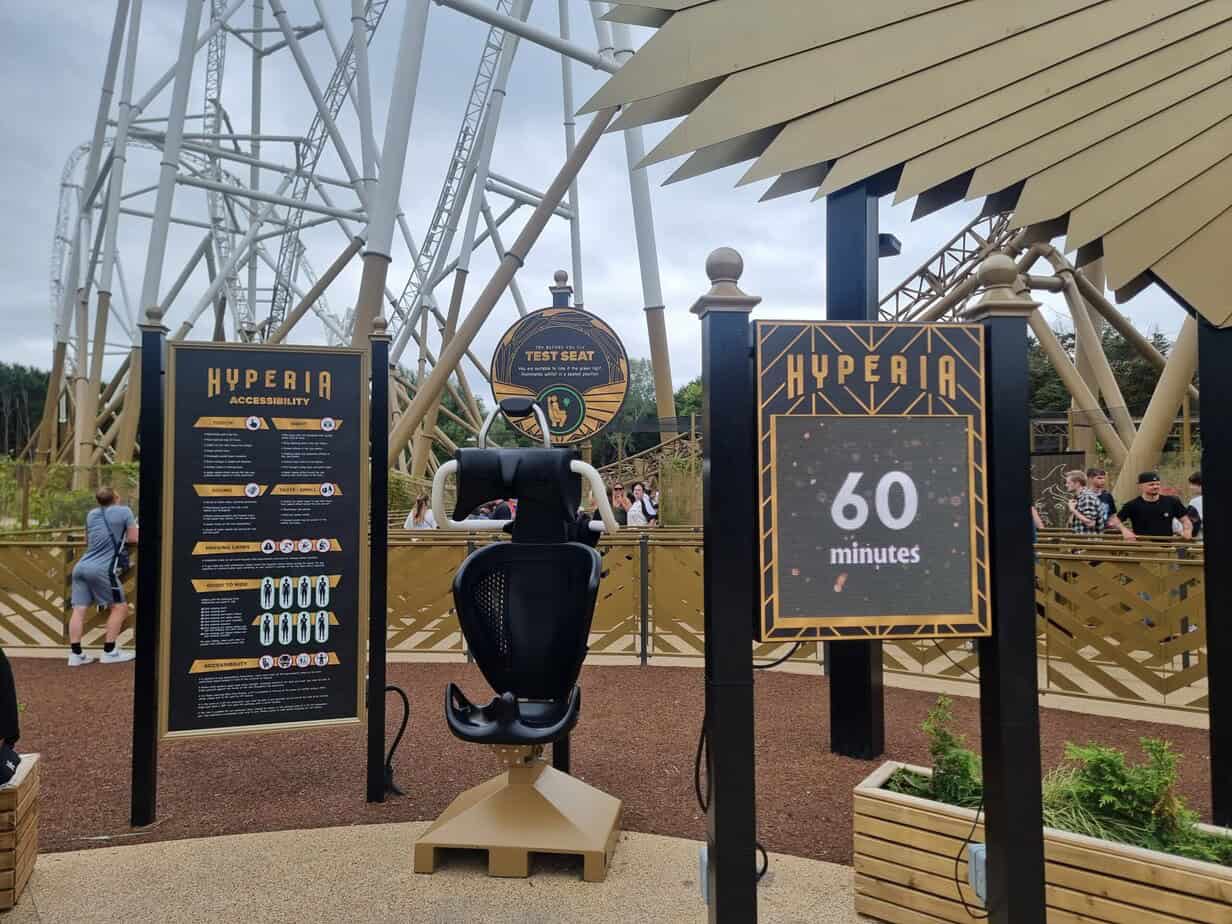 A black and gold-themed test seat for the Hyperia rollercoaster is displayed in front of detailed accessibility signage and a digital wait time board showing 60 minutes. This visual setup is part of ensuring inclusive ride experiences, especially for those navigating concerns about plus size at Thorpe Park.