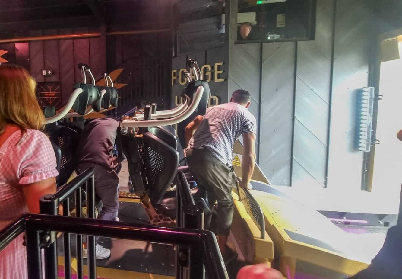 Guests prepare to board the Hyperia coaster as staff assist with harness checks in a dark, futuristic loading bay marked with “FORGE YARD” signage.