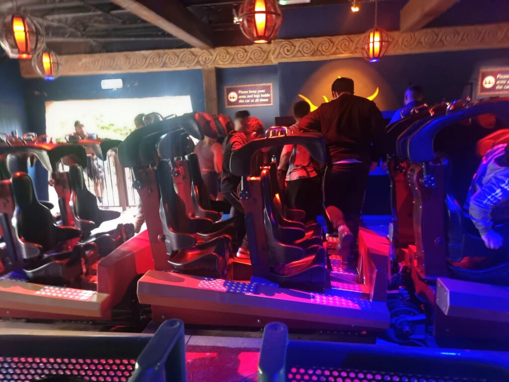 Guests are seen boarding a rollercoaster in a dimly lit indoor loading bay, with overhead signs and moody lighting enhancing the immersive ride experience.