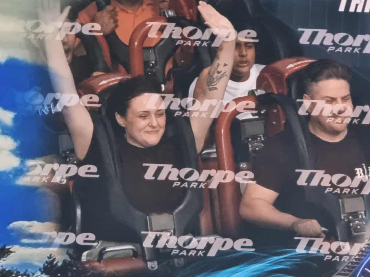 A close-up on-ride photo shows three people strapped into a rollercoaster seat with harnesses, smiling and screaming mid-ride with “Thorpe Park” watermarks layered over the image.