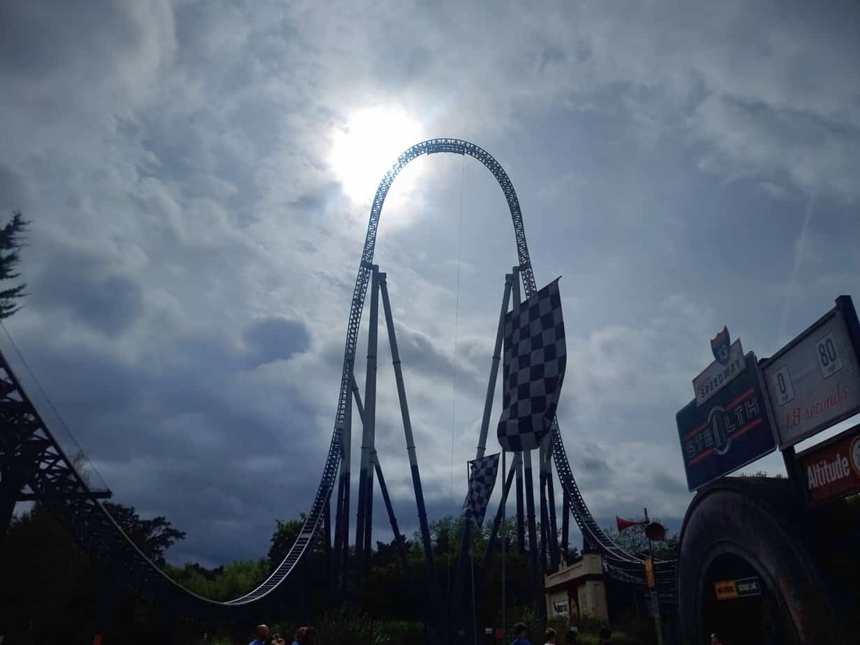 The towering top hat element of the Stealth rollercoaster stretches into a sunlit sky, with checkered flags and a sign showing “0 to 80 in 1.8 seconds” emphasizing the ride’s speed.